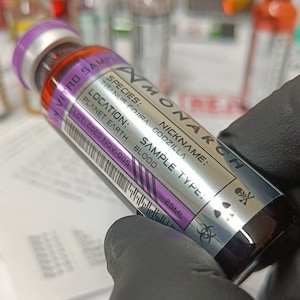 Puede incluir: Un vial plateado etiquetado como "MONARCH" con un símbolo de calavera y huesos cruzados. El vial contiene una muestra de sangre de un Titanus Gojira Godzilla, etiquetado como "TIPO DE MUESTRA: SANGRE".