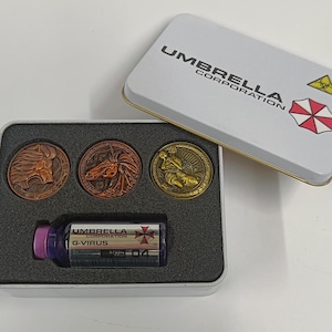 Op de afbeelding: Een witte blikken doos met de woorden "Umbrella Corporation" gedrukt op de deksel. De doos bevat drie gouden munten met verschillende ontwerpen en een klein glazen flesje met een paarse dop met het label "Umbrella Corporation G-Virus".