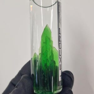以下が含まれることがあります： 銀色の蓋が付いた透明なガラス瓶に、緑色の結晶状物質が入っています。瓶には「LEXCORP」と「Atoms #119 -  Crystalline Potassium」と書かれたラベルが付いています。