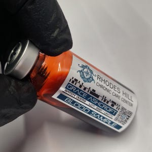 May include: A vial filled with an orange liquid, labelled "BLOOD SAMPLE" with the text "RHODES HILL CHRONIC CARE CENTER" and "GRACE ASHCROFT". The vial is held by a gloved hand.