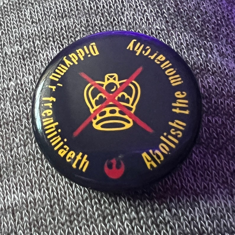 Pode incluir: Um bot&atilde;o preto com um X vermelho sobre uma coroa dourada e o texto "Abolish the Monarchy" em branco. O texto "freihini&aelig;eth" est&aacute; escrito em um c&iacute;rculo ao redor da coroa e o texto "K43jeuwon" est&aacute; escrito em um c&iacute;rculo ao redor do texto "Abolish the Monarchy".