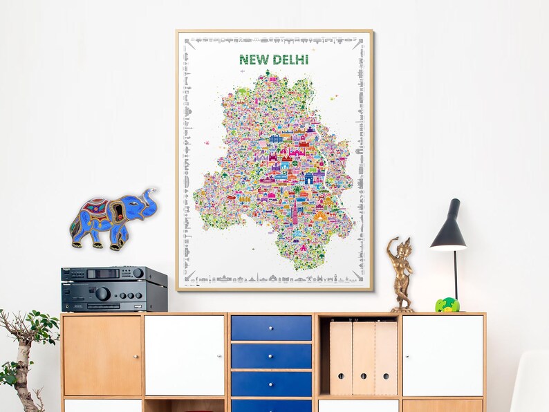 A vibrant, illustrated map of New Delhi, India, features numerous colorful landmarks and buildings. The map is framed and displayed on a wall above a light wood cabinet with blue drawers.  A small, blue, decorative elephant and a gold statue are also visible. The map's title, 'NEW DELHI,' is prominently displayed at the top. The overall aesthetic is modern and eclectic, blending cultural elements with contemporary design.