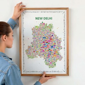 Person hanging a framed colorful New Delhi illustrated map poster in a natural wood frame on a light wall.
