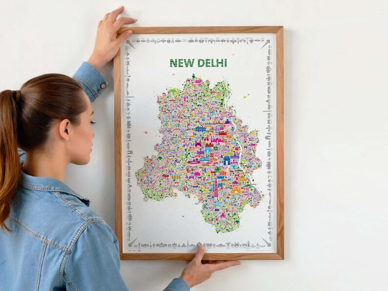 Person hanging a framed colorful New Delhi illustrated map poster in a natural wood frame on a light wall.