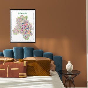 A framed wall art print featuring a colorful, detailed map of New Delhi, India. The map is filled with various small, vibrant icon illustrations of landmarks and cultural icons, set against a white background with a black frame. It is an artistic artwork.