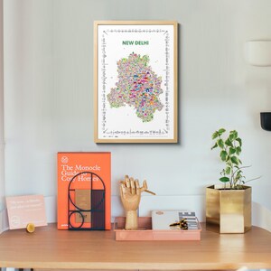 A framed vertical wall art print featuring a colorful illustrated map of New Delhi, India. The map poster is centered on a white background with the text NEW DELHI in green at the top and a border of small black and white landmark icons.