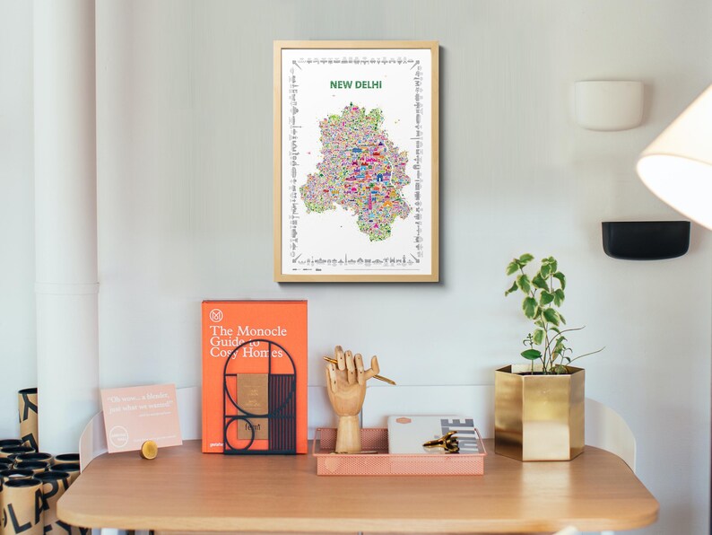 A framed vertical wall art print featuring a colorful illustrated map of New Delhi, India. The map poster is centered on a white background with the text NEW DELHI in green at the top and a border of small black and white landmark icons.