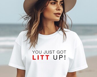 May include: White t-shirt with the text "YOU JUST GOT LITT UP!" in black and red lettering. The shirt is being worn by a person with long brown hair and a white hat.