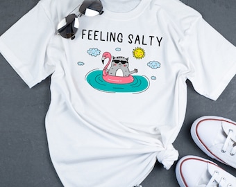 May include: White t-shirt with the text "Feeling Salty" and a cartoon cat wearing sunglasses and riding a pink flamingo float in blue water.