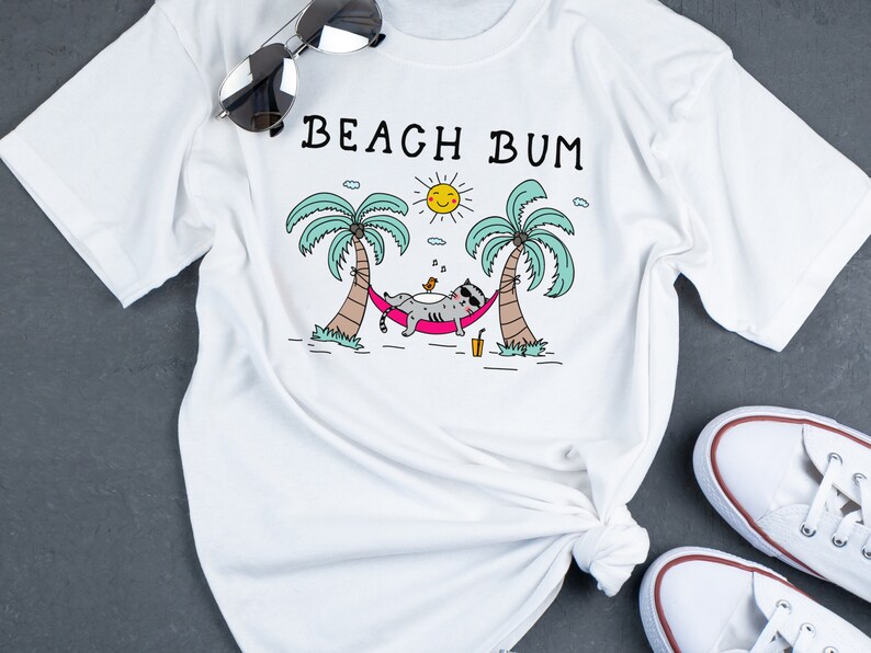 May include: White t-shirt with a graphic of a cat relaxing in a hammock between two palm trees. The text "Beach Bum" is printed above the graphic.