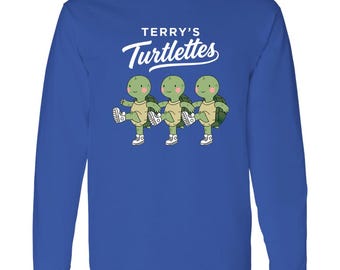 Könnte beinhalten: Ein königsblaues Langarmshirt mit einer Grafik von drei grünen Schildkröten, die weiße Turnschuhe tragen und einen Kick-Tanz aufführen. Über den Schildkröten steht in weißer Schrift TERRY'S Turtlettes, wobei das zweite Wort in Schreibschrift gehalten ist.