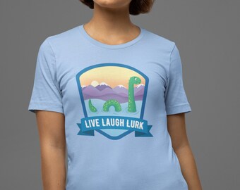 May include: Light blue t-shirt with a graphic of a green Loch Ness Monster in a lake with mountains in the background. The graphic is surrounded by a blue and yellow shield with the text "LIVE LAUGH LURK" on a white banner.