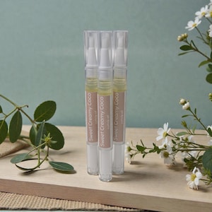 May include: Three cuticle oil pens labelled "Sweet Creamy Coco" on a wooden surface. The pens are clear with white and tan labels. The background is a light green colour with white flowers and green leaves.