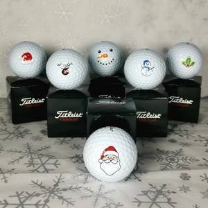 May include: A selection of Titleist golf balls with Christmas-themed designs. Each white ball has a different festive graphic, such as Santa Claus, a snowman, and holly. The golf balls are presented on top of black boxes.