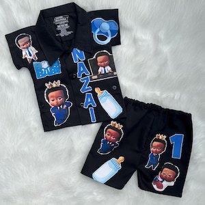 May include: Black baby outfit set featuring a button-down shirt and shorts. The shirt has "Boss Baby" and "Nazai" text, baby bottle and pacifier graphics. The shorts have the number "1" and baby graphics. The outfit is decorated with baby-themed graphics.