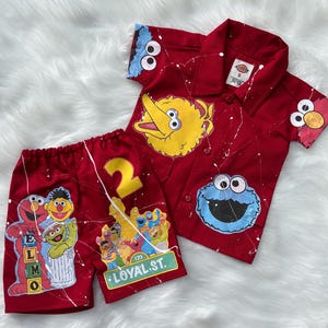 Op de afbeelding: Rode korte mouwen shirt en short set met Sesamstraat-personages. Het shirt heeft Big Bird, Cookie Monster en Elmo graphics, met witte verf spatter details. De shorts hebben het nummer 2 en andere Sesamstraat-personages. De shorts hebben ook de tekst "Loyal St."