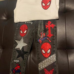 Könnte beinhalten: Ein Set aus weißem T-Shirt und schwarzer Jeans. Das T-Shirt hat einen roten Spider-Man-Maskenaufnäher. Die Jeans hat mehrere Aufnäher, darunter Spider-Man-Masken, Sterne und die Wörter "groovy" und "nope". Eine silberne Kette ist an der Jeans befestigt.