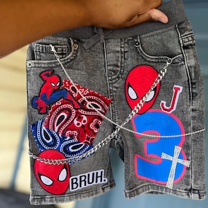 Puede incluir: Pantalones cortos vaqueros grises con parches de Spiderman, un diseño de cachemira rojo y azul, y el número "3" en azul. Los pantalones cortos tienen un detalle de cadena plateada y la palabra "BRUH" está impresa en la parte delantera.