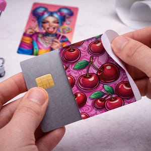 May include: A gray credit card with a gold chip is being peeled to reveal a cherry design. The cherry design features red cherries with green leaves on a pink background. A colorful card with a cartoon image is in the background.