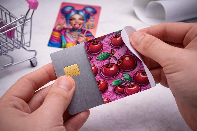 May include: A gray credit card with a gold chip is being peeled to reveal a cherry design. The cherry design features red cherries with green leaves on a pink background. A colorful card with a cartoon image is in the background.