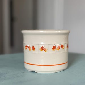 May include: A cream-colored ceramic crock featuring a repeating pattern of orange, yellow, and white candy corn motifs around the center. The cylindrical container has a textured basket-weave band and a single thin orange horizontal line near the base.