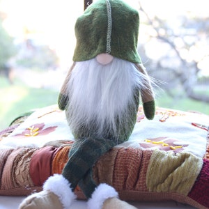 May include: A green gnome with a white beard sits on a multi-colored patchwork pillow. The pillow has a brown, orange, and red color scheme.