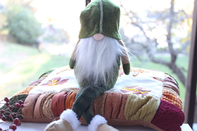 May include: A green gnome with a white beard sits on a multi-colored patchwork pillow. The pillow has a brown, orange, and red color scheme.