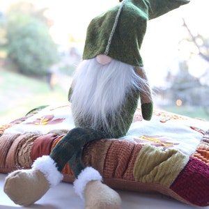 May include: A green gnome with a white beard sits on a colorful patchwork pillow. The gnome is wearing a green hat and has brown boots.