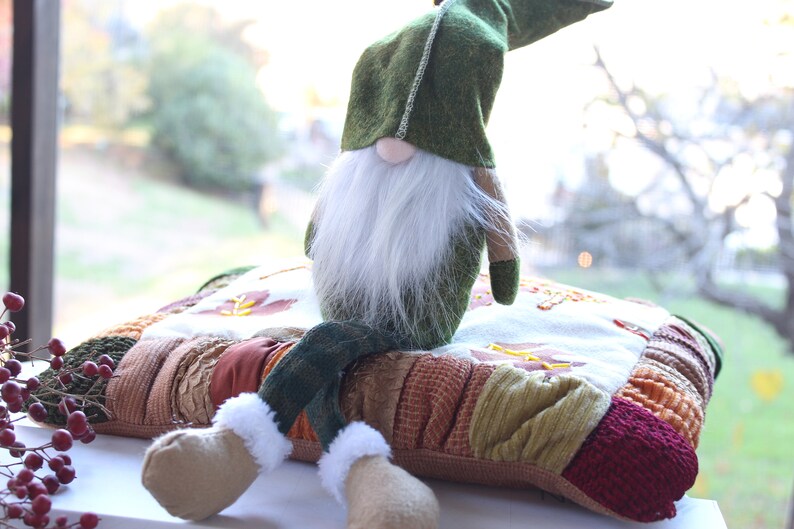 May include: A green gnome with a white beard sits on a colorful patchwork pillow. The gnome is wearing a green hat and has brown boots.
