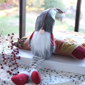 May include: A gray and red gnome with a white beard sits on a multi-colored patchwork pillow. The gnome has a pointed hat and striped legs. The gnome is sitting on a white box on a table with a speckled surface.