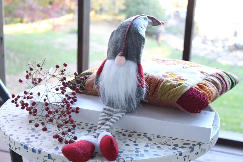 May include: A gray and red gnome with a white beard sits on a multi-colored patchwork pillow. The gnome has a pointed hat and striped legs. The gnome is sitting on a white box on a table with a speckled surface.