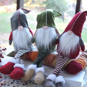 May include: Three gnomes with long legs and white beards are sitting on a colorful patterned cushion. The gnomes are wearing red, green, and gray hats.