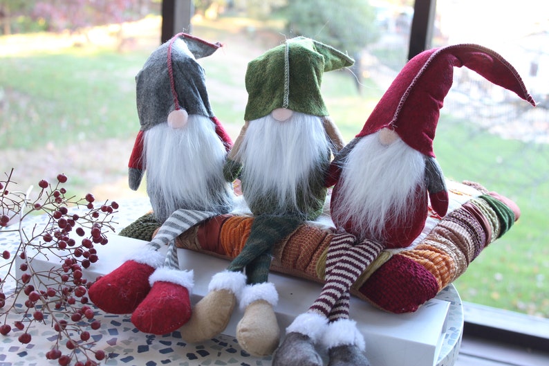 May include: Three gnomes with long legs and white beards are sitting on a colorful patterned cushion. The gnomes are wearing red, green, and gray hats.