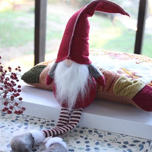 May include: A red and white striped gnome with a long white beard sits on a multi-colored patchwork pillow. The gnome is wearing a red hat with a white trim and gray mittens.