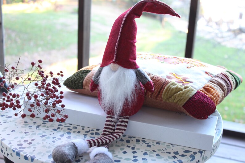May include: A red and white striped gnome with a long white beard sits on a multi-colored patchwork pillow. The gnome is wearing a red hat with a white trim and gray mittens.