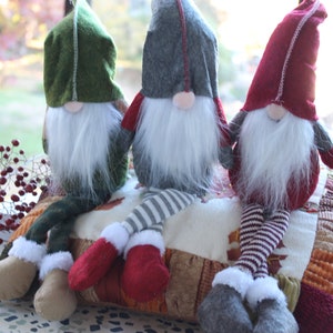 May include: Three gnomes sitting on a multi-colored patchwork cushion. The gnomes are wearing red, green, and gray hats with white fur trim. They have white beards and are wearing striped pants.
