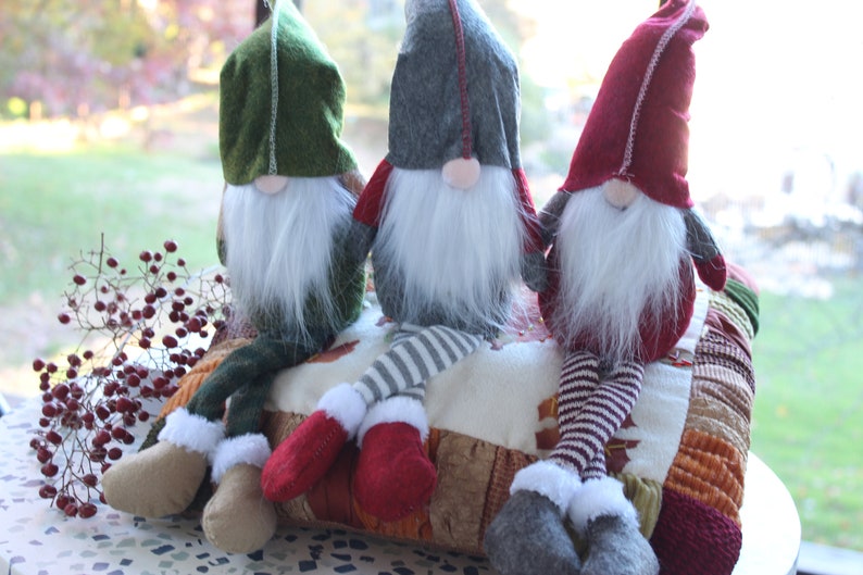 May include: Three gnomes sitting on a multi-colored patchwork cushion. The gnomes are wearing red, green, and gray hats with white fur trim. They have white beards and are wearing striped pants.