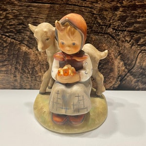 May include: A porcelain figurine of a young girl in a red and brown dress holding a basket of flowers. A white goat sits beside her. The figurine is on a green base.