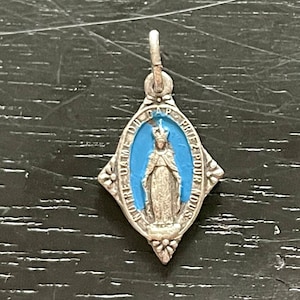 May include: A silver pendant with a blue enamel oval featuring a depiction of the Virgin Mary. The oval is surrounded by a diamond-shaped frame with floral accents. The text "NOTRE DAME DU GAP PRIEZ POUR NOUS" is inscribed around the oval.