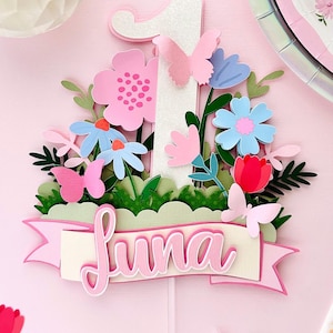 May include: A pink and white cake topper with a glittery number one, flowers, butterflies, and a banner that says "Luna".