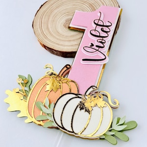 May include: A pink number one cake topper with the name "Violet" written on it. The topper is decorated with two pumpkins, one orange and one white, and gold leaves.