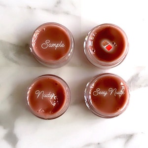May include: Four small jars of lip gloss with different shades of brown. The jars are labeled "Sample", "Nude", "Sexy Nude", and one jar has a small chocolate bar sticker.