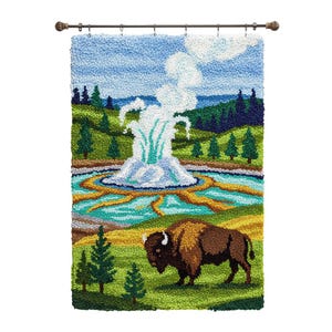 May include: A handcrafted wall hanging depicting a geyser erupting in a colorful landscape. The artwork features a bison in a grassy field, with a blue sky and green trees. The piece is mounted on a decorative rod.