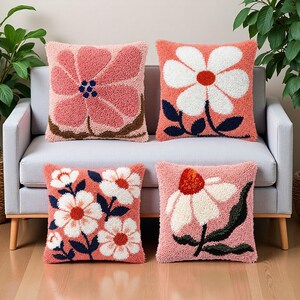 May include: Four square throw pillows with floral designs. The pillows feature a textured, tufted appearance with a coral pink background. The flowers are white, coral, and pink with dark blue and brown accents. The pillows are on a gray sofa.