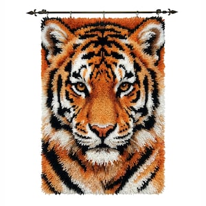 May include: A detailed, textured wall hanging featuring a tiger's face. The artwork uses orange, black, and white fibers to create the tiger's fur and facial features. The piece is mounted on a decorative rod.
