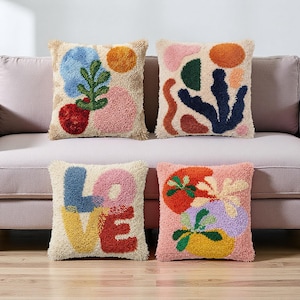 May include: Four square decorative throw pillows with abstract designs. Each pillow features a cream-colored background with colorful, textured embroidery. One pillow spells "LOVE" in blue, pink, yellow, and red. The other three have floral and geometric patterns.