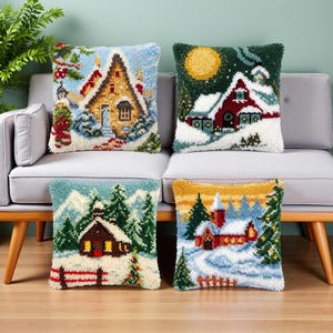 Peut inclure: Quatre coussins carrés présentant des motifs en punch needle texturé de scènes hivernales. Chaque coussin affiche une maison ou un chalet différent entouré de neige, de sapins et d'éléments festifs dans des tons de rouge, vert, jaune et blanc.