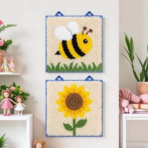 May include: Two square wall hangings with a blue border. One features a yellow and black bee, the other a yellow sunflower with a brown center. Both are on a cream background with green grass.