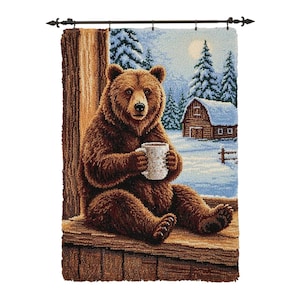 Könnte beinhalten: Ein brauner Bär sitzt auf einer Holzplattform und hält einen weißen Becher. Der Bär ist der Mittelpunkt eines Wandteppichs, der eine verschneite Winterlandschaft mit einer Hütte und Kiefern zeigt. Der Wandteppich hängt an einer dekorativen schwarzen Stange.