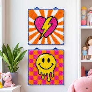 Könnte beinhalten: Zwei quadratische Wandbehänge mit blauen Rändern. Eines zeigt ein rosa Herz mit einem gelben Blitz vor einem orange-weißen Hintergrund. Das andere hat ein gelbes, schmelzendes Smiley-Gesicht auf einem rosa-orangenen Schachbrettmuster.